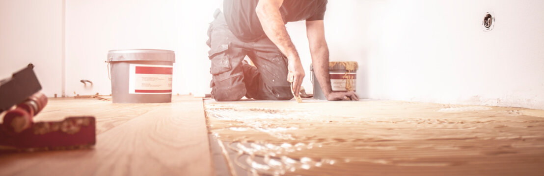 Handwerker verlegt Parkettboden und streicht den Estrich mit Kleber ein und schl&auml;gt die Dielen mit dem Hammer und dem Schlagklotz zusammen