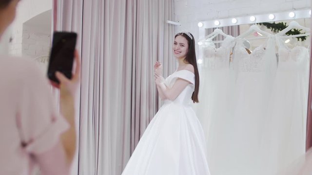 Rear view of female saleswoman making photos of joyful woman wearing wonderful wedding dress using phone camera