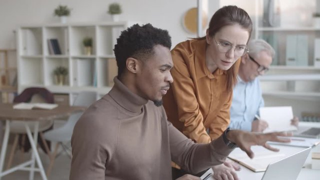 Medium arc shot of Caucasian female and African American colleagues looking at laptop screen together, discussing work project, talking, thinking and tentatively proposing different ideas