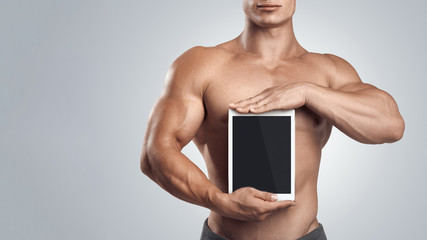 Close up photo of a shirtless young fitness male model holding vertically digital tablet with blank screen, isolated on white background.