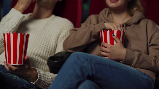 Medium shot of unrecognizable young couple eating popcorn and talking while watching film in cinema