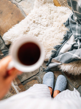 Top view of a pair of gray home slippers on female's feet near the natural white sheep sheepskin with warm plaid dropped on the stone floor in the cozy bedroom with unfocused hand with cup of coffee.