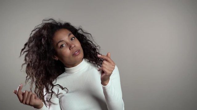 African american woman with curly hair having fun, smiling, dancing with head and snaps fingers in studio against gray background. Music, dance concept, slow motion.