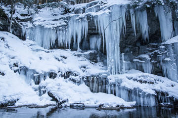 Winter frozen waterfall. Small pond and snowy boulders bellow cascade of waterfall. Crystal freeze...