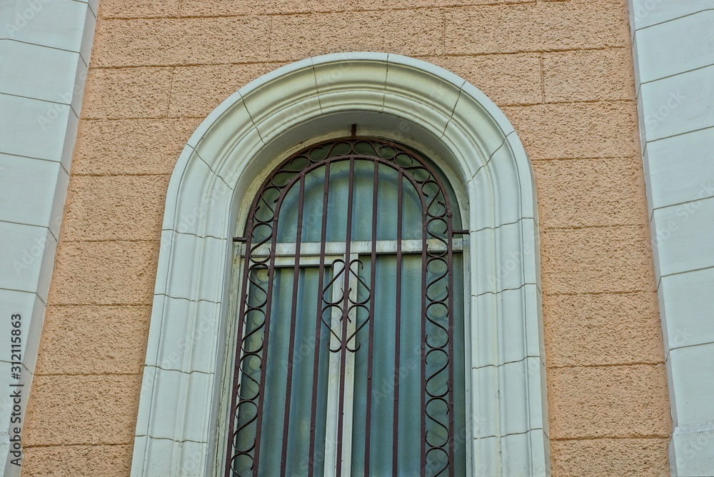 Sticker one large gray old window behind the iron bars against the brown concrete wall of the building