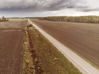 Small road in a country side by ploughed agricultural fields. Cloudy sky, Selective focus, Forest in the background. Aerial  view.