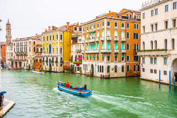 Boats and water taxis sailing and docked on Grand Canal along striped and wooden mooring poles and colorful Venetian architecture buildings. People/ tourists in Venice city Italy on rainy day.
