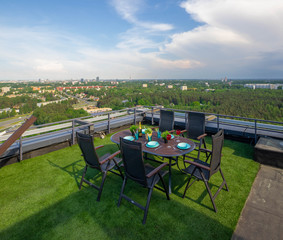 Rooftop terrace with green grass. Laid table with chairs. View of the city from the top.