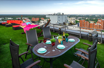 Laid table, cozy couch and hammock on the rooftop terrace with green grass. Summer day.