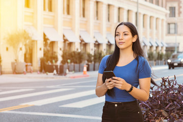 Young attractive girl awaits a ride share car in the city - Holding smart phone waiting on the sidewalk - Daytime with warm lighting