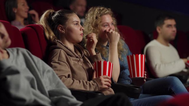Side view medium shot of two cheerful young women eating popcorn and talking while watching movie sitting on comfortable seats in cinema
