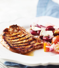Grilled pork steak with Kidney bean, cherry tomatoes, onion and feta cheese on bright wooden background. Close up. 