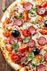 Pizza with sausage, cherry tomatoes, black olives and fresh herbs. Home made food. Concept for a tasty and hearty meal. Brown wooden background. Top view. 