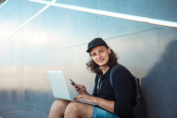 A young man, sitting on a skateboard, is trying to synchronize a laptop with a phone. Look into the...