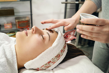 Obraz na płótnie Canvas Young woman coffee face treatment at spa salon cosmetologist applies mask for peeling on the face of a girl in a cosmetology room cosmetic therapy lying on bed covered with towel