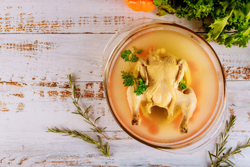 Chicken broth with carrots, whole chicken, onion, celery and parsley in glass pot.