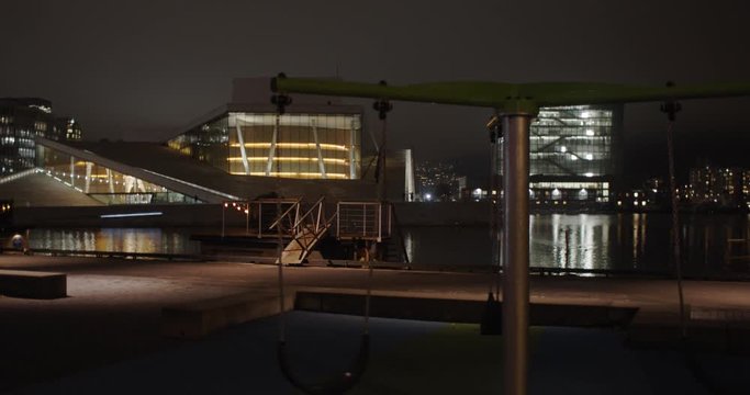 Medium full 4K professional shot with slow parallax motion of empty swings in playground with the illuminated Opera Music House and Edvard Munch gallery Lambda in background, at night in Oslo Norway.