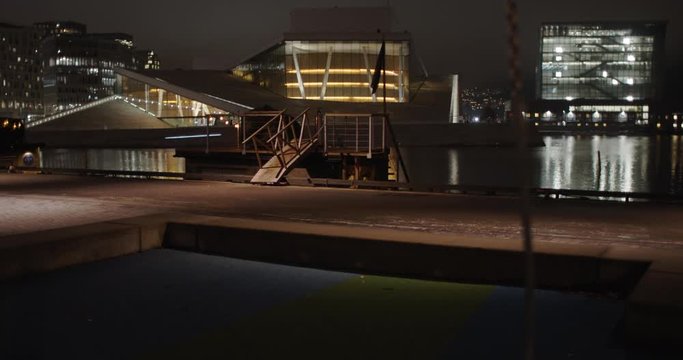 Medium full 4K shot with slow forward tilt reveal motion of playground with empty swings and illuminated Opera Music House and Edvard Munch Museum Lambda in background, at winter night in Oslo Norway.
