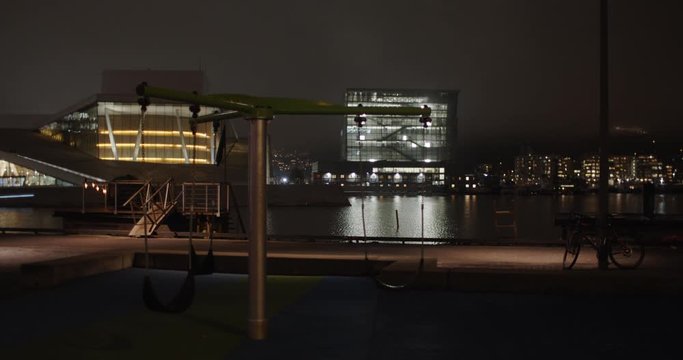 Medium full 4K professional shot with slow parallax motion of empty swings in playground with the illuminated Opera Ballet House and Edvard Munch museum Lambda in background, at night in Oslo Norway.