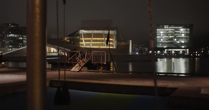 Medium full 4K shot with slow forward tilt reveal motion of playground with empty swings and illuminated Opera House Theatre and Edvard Munch gallery Lambda in background, at night in Oslo Norway.