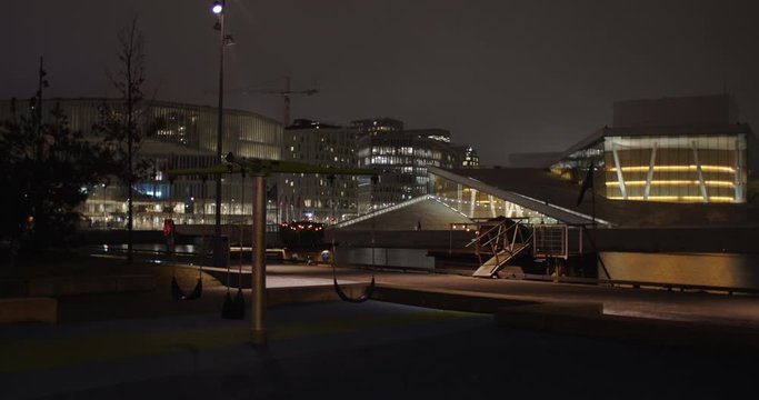 Medium full professional cinema 4K shot with parallax motion of empty swings on playground in front of illuminated Oslo Opera Theatre and Edvard Munch Gallery Lambda, at night in Bj&oslash;rvika Oslo Norway.
