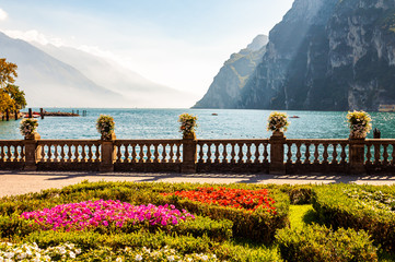 Garda lake promenade with colorful flowerbeds with growing and blooming plants, classic stone fence built on the edge with flowerpots with blooming flowers. Garda lake and mountains on background