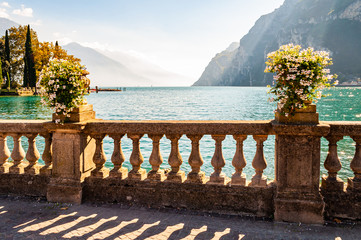 Beautiful Garda lake promenade with classic stone fence railings built on the edge with flowerpots with blooming white flowers. Garda lake surrounded by the high dolomite mountains on the background