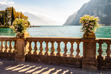 Beautiful Garda lake promenade with classic stone fence railings built on the edge with flowerpots with blooming white flowers. Garda lake surrounded by the high dolomite mountains on the background