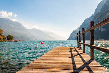 Scenic view on wooden planks pier with railings built on northern shore of beautiful Garda lake in Lombardy, Italy surrounded by high dolomite mountains and crystal clear blue water of the lake. Riva