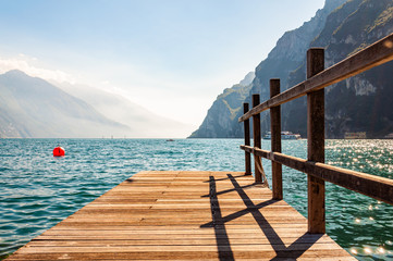 Scenic view on wooden planks pier with railings built on northern shore of beautiful Garda lake in Lombardy, Italy surrounded by high dolomite mountains and crystal clear blue water of the lake. Riva