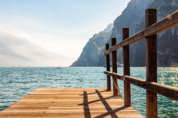 Scenic view on wooden planks pier with railings built on northern shore of beautiful Garda lake in Lombardy, Italy surrounded by high dolomite mountains and crystal clear blue water of the lake. Riva