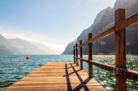 Scenic view on wooden planks pier with railings built on northern shore of beautiful Garda lake in Lombardy, Italy surrounded by high dolomite mountains and crystal clear blue water of the lake. Riva