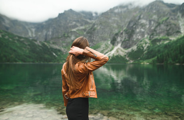 Stylish girl in casual casual clothes stands on the shore of a mountain lake and looks at the...