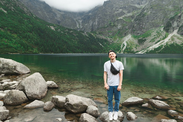 Smiling young man in a cap and casual clothes stands against the backdrop of Lake Morskie Oko in the Tatra Mountains, looks into the camera and smiles. Happy hiker hiking lake in mountains