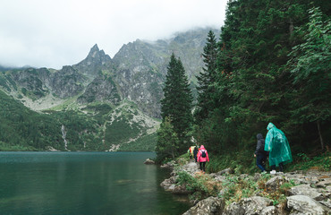 Group of tourists in raincoats walk along a rock path near beautiful Morskie Oko Lake with clear water against a rocky Tatra Mountains background. Tourists and road hiking in the rain. © bodnarphoto