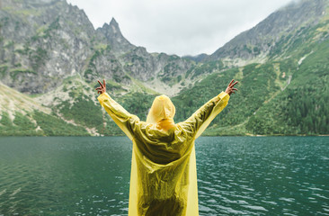 Joyful person in yellow raincoat with raised hands stands against unreal mountain landscape with lake. Person hiking on Lake Morskie Oko in Tatra National Park. Background. Copy space © bodnarphoto