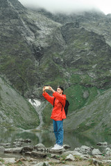 Portrait of young man in red raincoat and jacket standing on mountains and lake background and taking photo on smartphone. Hiker man taking selfie in hiking mountains. Vertical photo.