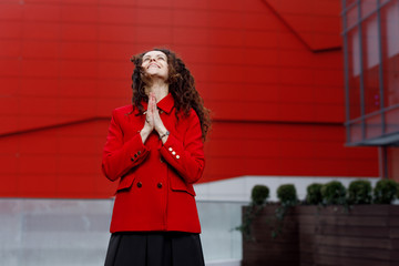 Happy young woman with curly hair dressed in red jacket stands with her hands clasped and face looking up. she is happy, is successful.