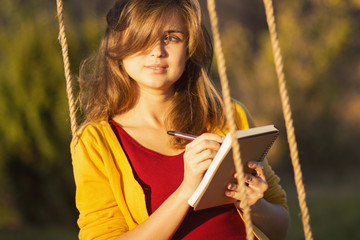 cute girl on a swing with a notebook and pen keeps a diary of feelings in nature, a woman composes...