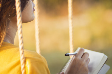cute girl on a swing with a notebook and pen keeps a diary of feelings in nature, a woman composes...