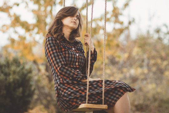 Upset Girl In A Dress Sways On A Swing In The Autumn Garden, A Young Woman In Melancholy Mood Thinkng On Nature