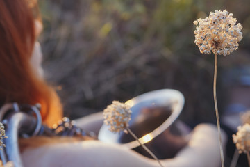 dreamy red-haired girl with saxophone on the shoulder sitting on the ground in a field among dried flowers and enjoys nature at sunset, young woman relaxing, concept of relax, lifestyle