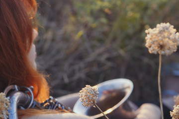 dreamy red-haired girl with saxophone on the shoulder sitting on the ground in a field among dried flowers and enjoys nature at sunset, young woman relaxing, concept of relax, lifestyle