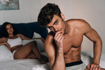 thoughtful man looking at camera while sitting on bed near sexy girlfriend