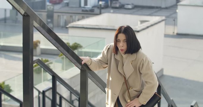 Portrait of exhausted Caucasian woman standing on stairs in the city and resting. Brunette businesswoman breathing heavily and walking up, late for meeting. Jobs, meetings. Cinema 4k ProRes HQ.