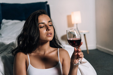 sensual, dreamy girl holding glass of red wine in bedroom