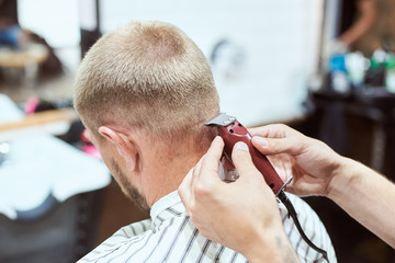 Dark blonde man at barber shop is having his nape shaved with a trimmer, hairdresser is holding a...