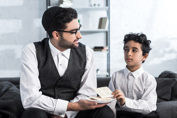 smiling jewish father holding plate and son taking matza in apartment