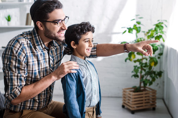 smiling jewish father pointing with finger and looking through window with son in apartment