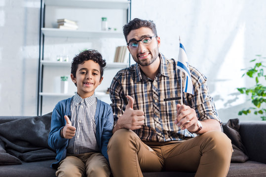 smiling jewish father with flag of israel and son showing thumbs up in apartment